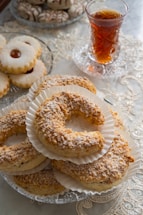 A table adorned with an assortment of pastries and a glass of tea. The pastries include crescent-shaped cookies dusted with powdered sugar and arranged on a decorative glass plate. In the background, jam-filled cookies and other delicacies are displayed. A glass of amber tea is placed nearby on a lace tablecloth, contributing to an elegant presentation.
