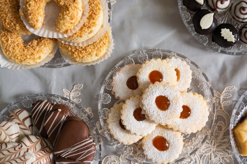 A variety of pastries and cookies are artistically arranged on decorative glass plates. There are jam-filled cookies dusted with powdered sugar, crescent-shaped pastries topped with nuts, heart-shaped chocolate-covered treats with white drizzle, and round chocolates adorned with white leaf patterns. The arrangement sits on an elegant lace tablecloth creating a refined and inviting display.