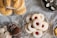 A variety of pastries and cookies are artistically arranged on decorative glass plates. There are jam-filled cookies dusted with powdered sugar, crescent-shaped pastries topped with nuts, heart-shaped chocolate-covered treats with white drizzle, and round chocolates adorned with white leaf patterns. The arrangement sits on an elegant lace tablecloth creating a refined and inviting display.