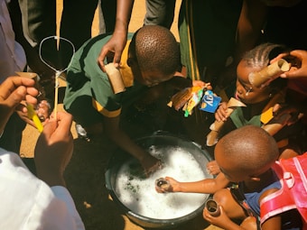 A group of children surround a basin filled with soapy water. They appear to be engaging in an outdoor activity, possibly blowing bubbles, as some of them hold circular bubble wands and cardboard tubes. Various hands are reaching into the frame, and the children are intently focused on the basin.