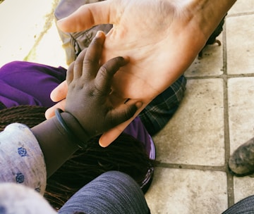 A close-up of two hands, one smaller and darker and the other larger and lighter, gently held together. The setting appears to be outdoors with a tiled ground visible beneath. The smaller hand seems to rest on the palm of the larger hand, conveying a sense of connection and care.