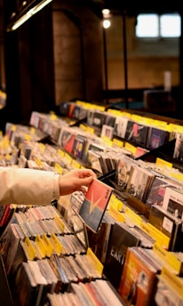 A person wearing a white jacket is browsing through a collection of CDs organized in rows on shelves at a music store. The CDs are arranged with labels on yellow tags for easy identification. The store has a warm, dimly lit atmosphere.