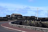 A rural scene featuring a paved road curving gently in the foreground, bordered by low stone walls and rocky terrain. A house with a brown roof and stone facade is situated at the center, surrounded by stone barriers. Telephone poles and wires are visible, with a bright blue sky and scattered clouds in the background.