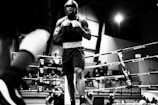 A boxer stands confidently in a ring, wearing a helmet and gloves. The scene captures the intensity of the moment, with ropes framing the area and spectators watching intently in the background. The photograph is in black and white, emphasizing the drama and athleticism.