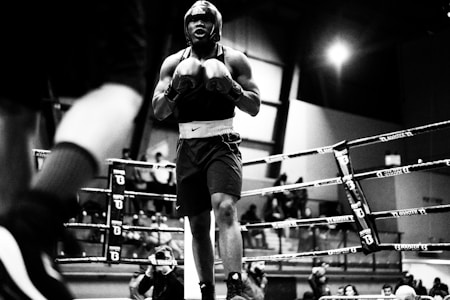 A boxer stands confidently in a ring, wearing a helmet and gloves. The scene captures the intensity of the moment, with ropes framing the area and spectators watching intently in the background. The photograph is in black and white, emphasizing the drama and athleticism.