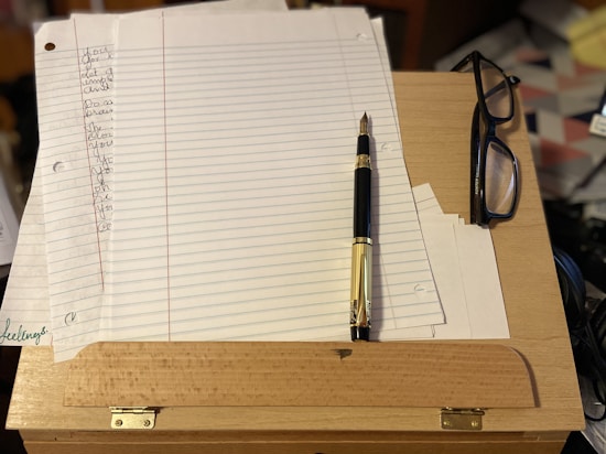 A wooden desk is topped with a pad of lined paper, a black and gold fountain pen, and a pair of glasses with black frames. Some of the paper contains handwritten text. The surface and surrounding area appear cluttered with various items partially visible in the background.