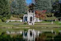 A serene mausoleum surrounded by lush greenery and a reflective pond. The structure is white with decorative statues and features, nestled amid towering trees and a vibrant red-orange tree. A bench sits on the grassy area, enhancing the calm atmosphere.