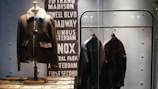 A display features three leather jackets. One is on a mannequin with a shearling collar, while two are hanging on a metal rack. The background includes a decorative panel with street names in bold capital letters.