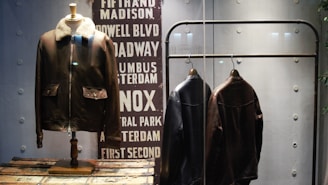 A display features three leather jackets. One is on a mannequin with a shearling collar, while two are hanging on a metal rack. The background includes a decorative panel with street names in bold capital letters.