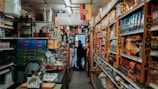 A narrow aisle in a small grocery store filled with various products from Colombia and Ecuador. The shelves are stocked with items such as packaged foods, beverages, and snacks. Signs and flags of Colombia and Ecuador are prominently displayed, adding a cultural touch to the setting. The lighting is warm and the store appears to be well-organized despite the tight space.