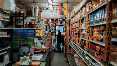 A narrow aisle in a small grocery store filled with various products from Colombia and Ecuador. The shelves are stocked with items such as packaged foods, beverages, and snacks. Signs and flags of Colombia and Ecuador are prominently displayed, adding a cultural touch to the setting. The lighting is warm and the store appears to be well-organized despite the tight space.