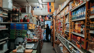 A narrow aisle in a small grocery store filled with various products from Colombia and Ecuador. The shelves are stocked with items such as packaged foods, beverages, and snacks. Signs and flags of Colombia and Ecuador are prominently displayed, adding a cultural touch to the setting. The lighting is warm and the store appears to be well-organized despite the tight space.