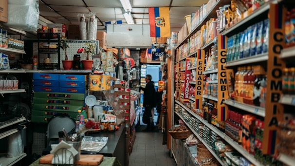 A narrow aisle in a small grocery store filled with various products from Colombia and Ecuador. The shelves are stocked with items such as packaged foods, beverages, and snacks. Signs and flags of Colombia and Ecuador are prominently displayed, adding a cultural touch to the setting. The lighting is warm and the store appears to be well-organized despite the tight space.