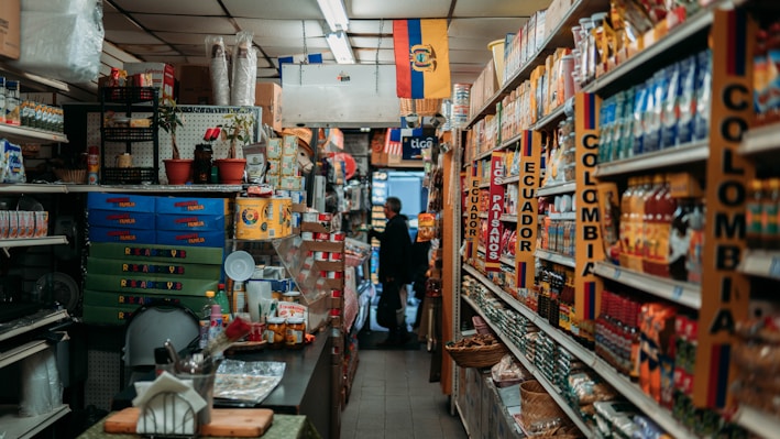 A narrow aisle in a small grocery store filled with various products from Colombia and Ecuador. The shelves are stocked with items such as packaged foods, beverages, and snacks. Signs and flags of Colombia and Ecuador are prominently displayed, adding a cultural touch to the setting. The lighting is warm and the store appears to be well-organized despite the tight space.