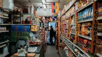 A narrow aisle in a small grocery store filled with various products from Colombia and Ecuador. The shelves are stocked with items such as packaged foods, beverages, and snacks. Signs and flags of Colombia and Ecuador are prominently displayed, adding a cultural touch to the setting. The lighting is warm and the store appears to be well-organized despite the tight space.