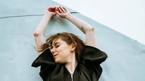 A person with long hair is stretching their arms upwards against a light blue, textured backdrop. The person is wearing a dark, loose-fitting top and appears to have a small tattoo on their forearm. Their eyes are closed, and they have a serene expression.