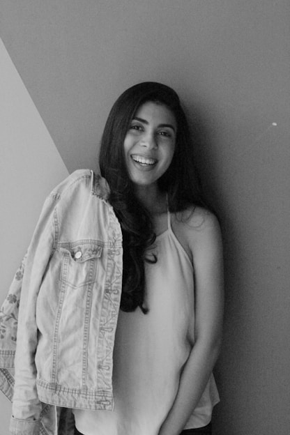 A young woman stands against a wall, smiling brightly at the camera. She has long, dark hair and is wearing a light-colored sleeveless top. A denim jacket is draped over her left shoulder.