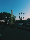 A tall vertical sign with 'HOLLYWOOD' written on it stands prominently against a clear blue sky. Several palm trees are visible in the background alongside urban buildings and parked cars. The scene captures a typical urban street setting during daytime.