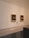 Two framed paintings hang on a large white wall in a gallery setting. The paintings appear to be classical portraits with dark backgrounds and figures highlighted in light. Below each painting are small, labeled plaques. The room has a polished stone floor and a minimalist interior.