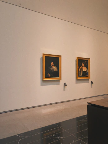 Two framed paintings hang on a large white wall in a gallery setting. The paintings appear to be classical portraits with dark backgrounds and figures highlighted in light. Below each painting are small, labeled plaques. The room has a polished stone floor and a minimalist interior.