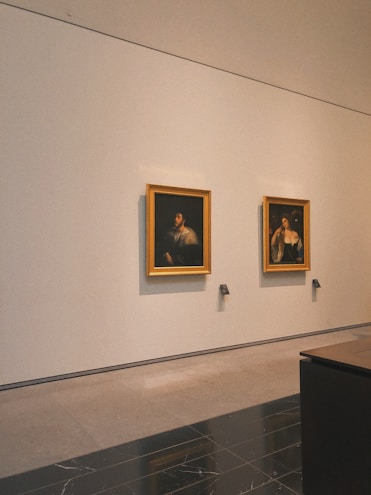 Two framed paintings hang on a large white wall in a gallery setting. The paintings appear to be classical portraits with dark backgrounds and figures highlighted in light. Below each painting are small, labeled plaques. The room has a polished stone floor and a minimalist interior.