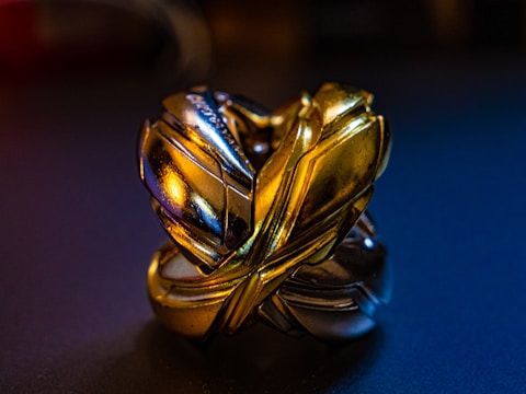 A metallic, intertwined puzzle sculpture with intricate design featuring interlocking pieces. The surface has a shiny finish with reflections of gold and silver hues.