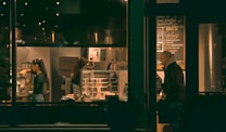A cozy, dimly lit cafe with warm lighting. People are working behind a counter, possibly preparing drinks. A menu board is visible on the wall, and shelves hold various items. There is a person seated near the window, seemingly engaged in conversation or reading.