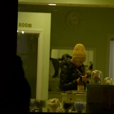 A person wearing a yellow knit hat and a dark puffy jacket is engrossed in their phone in a warmly lit indoor setting. They stand behind a counter with various objects, including a bottle and a dome covering a rose. The background shows a door marked with the word 'ROOM' and a wall clock.