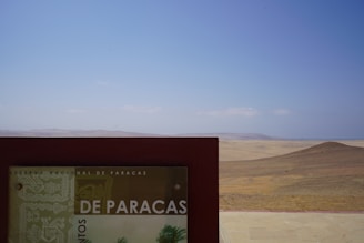 A scenic view of a desert landscape with a clear blue sky above. In the foreground, a sign with the text 'Reserva Nacional de Paracas' is partially visible, indicating this is a protected natural area.