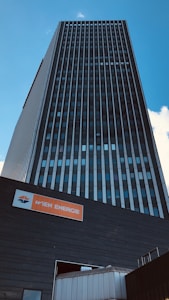 A tall, modern building with a dark facade and vertical lines extending upwards under a clear blue sky. At the base, there is a sign with the text 'WIEN ENERGIE' in white on an orange background.