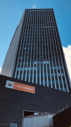 A tall, modern building with a dark facade and vertical lines extending upwards under a clear blue sky. At the base, there is a sign with the text 'WIEN ENERGIE' in white on an orange background.