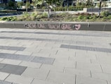 A modern outdoor area with a tiled walkway and landscaped greenery. On a concrete barrier, a message is written in pink chalk with the words 'EACH ONE TEACH ONE' and symbols including a peace sign and a heart.