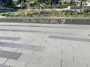 A modern outdoor area with a tiled walkway and landscaped greenery. On a concrete barrier, a message is written in pink chalk with the words 'EACH ONE TEACH ONE' and symbols including a peace sign and a heart.