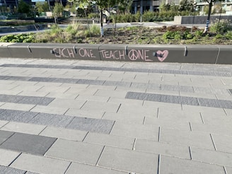 A modern outdoor area with a tiled walkway and landscaped greenery. On a concrete barrier, a message is written in pink chalk with the words 'EACH ONE TEACH ONE' and symbols including a peace sign and a heart.