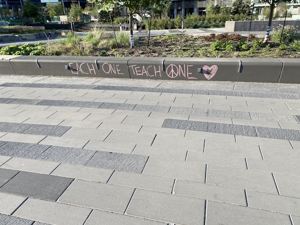 A modern outdoor area with a tiled walkway and landscaped greenery. On a concrete barrier, a message is written in pink chalk with the words 'EACH ONE TEACH ONE' and symbols including a peace sign and a heart.
