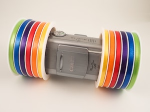 A silver Canon PowerShot S45 camera is positioned horizontally, with several colorful rolls of tape placed on either side of the camera. The tapes are arranged in an orderly fashion with the colors including green, blue, purple, red, orange, and yellow. The tapes provide a vibrant contrast to the metallic finish of the camera.