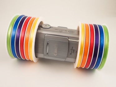 A silver Canon PowerShot S45 camera is positioned horizontally, with several colorful rolls of tape placed on either side of the camera. The tapes are arranged in an orderly fashion with the colors including green, blue, purple, red, orange, and yellow. The tapes provide a vibrant contrast to the metallic finish of the camera.