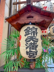 A traditional Japanese lantern with black kanji characters is surrounded by palm leaves. The decorative wooden frame above the lantern adds an authentic touch, and additional lanterns can be seen in the background.