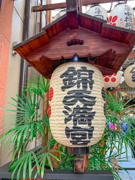 A traditional Japanese lantern with black kanji characters is surrounded by palm leaves. The decorative wooden frame above the lantern adds an authentic touch, and additional lanterns can be seen in the background.