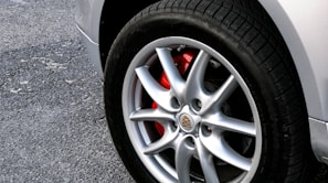 A close-up view of a car wheel with a shiny silver alloy rim. The brake caliper behind the rim is painted red, adding a contrasting color. The tire has a black tread and is mounted on a light-colored, slightly worn pavement.