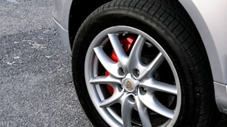 A close-up view of a car wheel with a shiny silver alloy rim. The brake caliper behind the rim is painted red, adding a contrasting color. The tire has a black tread and is mounted on a light-colored, slightly worn pavement.