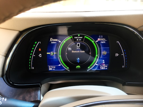 A car dashboard display showing various metrics including battery level, fuel range, odometer reading, and Bluetooth audio connectivity. The display includes indicators for power usage in kilowatts, EV range in miles, and a speedometer set to zero mph. The dashboard has a sleek, modern design with vibrant colors.