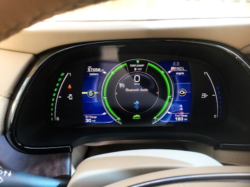 A car dashboard display showing various metrics including battery level, fuel range, odometer reading, and Bluetooth audio connectivity. The display includes indicators for power usage in kilowatts, EV range in miles, and a speedometer set to zero mph. The dashboard has a sleek, modern design with vibrant colors.
