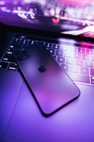 A smartphone with a sleek design is resting on the keyboard of a laptop. The lighting is a combination of vibrant purples and pinks, creating a futuristic and tech-savvy atmosphere.