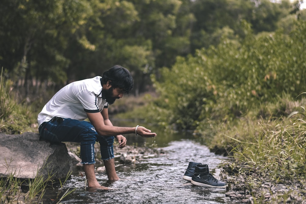 man in white t-shirt and blue denim jeans sitting on rock in river during daytime