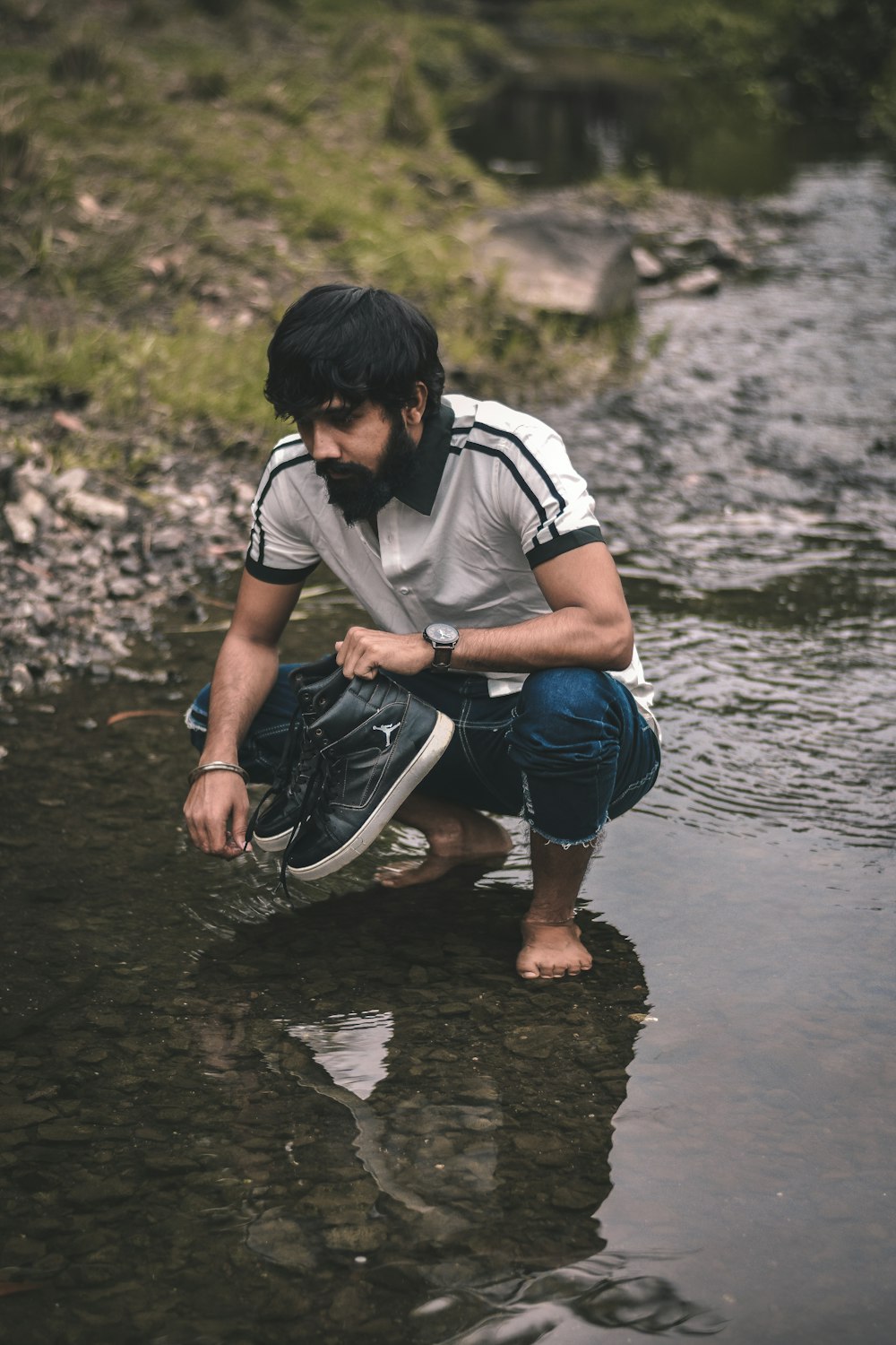 man in white and black shirt and blue denim jeans sitting on rock in water