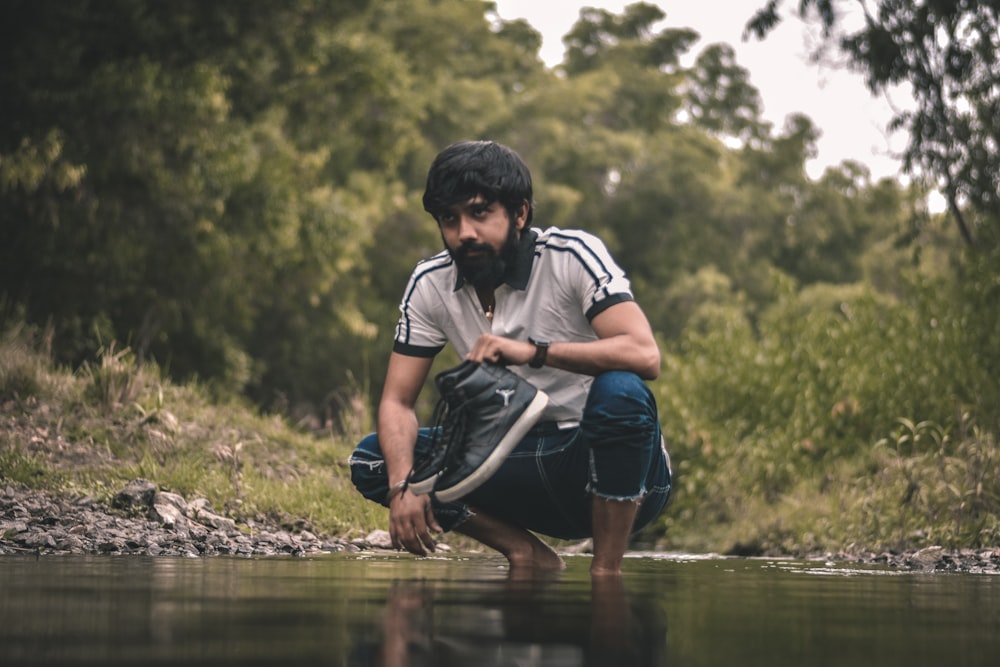 Mann in weiß-schwarzem T-Shirt und blauen Jeansshorts sitzt auf Felsen in der Nähe des Flusses