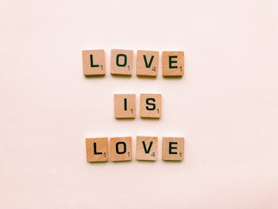 Wooden Scrabble tiles spell out the phrase 'LOVE IS LOVE' on a light beige background. The tiles are neatly aligned in three rows with letters and number values visible on each tile.
