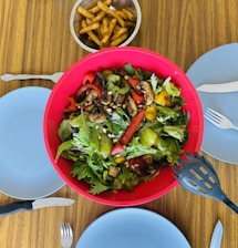 A vibrant salad with mixed greens, bell peppers, mushrooms, and pine nuts is placed in a large red bowl. Beside the salad, there is a small bowl of golden-brown French fries. The table is set with four blue plates, each accompanied by a fork and knife. The setting suggests preparation for a shared meal.