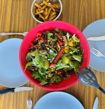 A vibrant salad with mixed greens, bell peppers, mushrooms, and pine nuts is placed in a large red bowl. Beside the salad, there is a small bowl of golden-brown French fries. The table is set with four blue plates, each accompanied by a fork and knife. The setting suggests preparation for a shared meal.